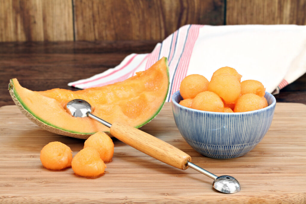 melon baller on a slice of melon with melon balls beside it and a bowl of melon bowls beside it